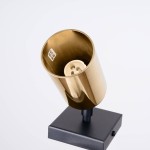 Kinkiet Winx w kolorze czarnym i złotym, z metalowym kloszem w formie cylindra. Widoczna oprawka GU10 w środku. Ustawiony na prostokątnej podstawie w kolorze czarnym, zdjęcie z góry.