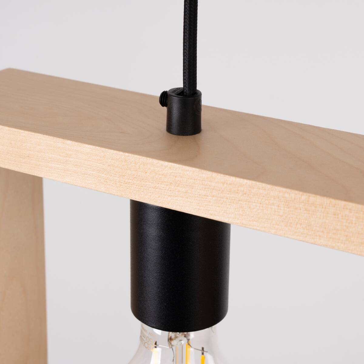 Lampa wisząca Aries 1xE27 w kolorze czarnym z drewnianym elementem. Widoczny metalowy uchwyt z czarnym wykończeniem i przezroczysta żarówka. Kąt ujęcia z góry, pokazujący detal mocowania i fakturę drewna.