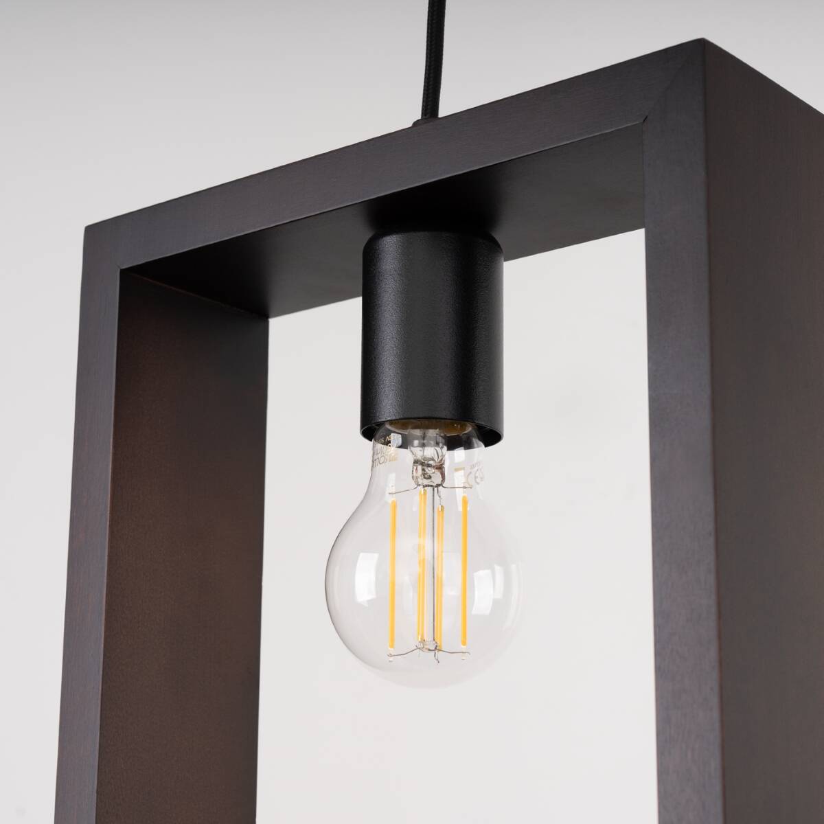 Lampa wisząca Aries 1xE27 w kolorze czarnym i wenge. Posiada prostokątną ramę z drewna wenge, z czarnym uchwytem dla żarówki. Widoczna przezroczysta żarówka z żółtymi włóknami. Ujęcie z bliska na detal lampy.