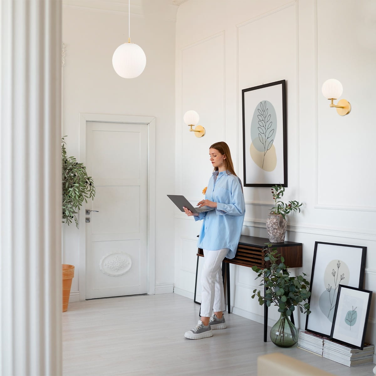 Kinkiet Paloma 1xE14 w mosiężnej oprawie z białym kloszem, zamontowany na ścianie. W tle widoczna roślinność oraz grafiki na ścianie, co tworzy nowoczesne wnętrze. Produkt o eleganckim designie, idealny do aranżacji przestrzeni.