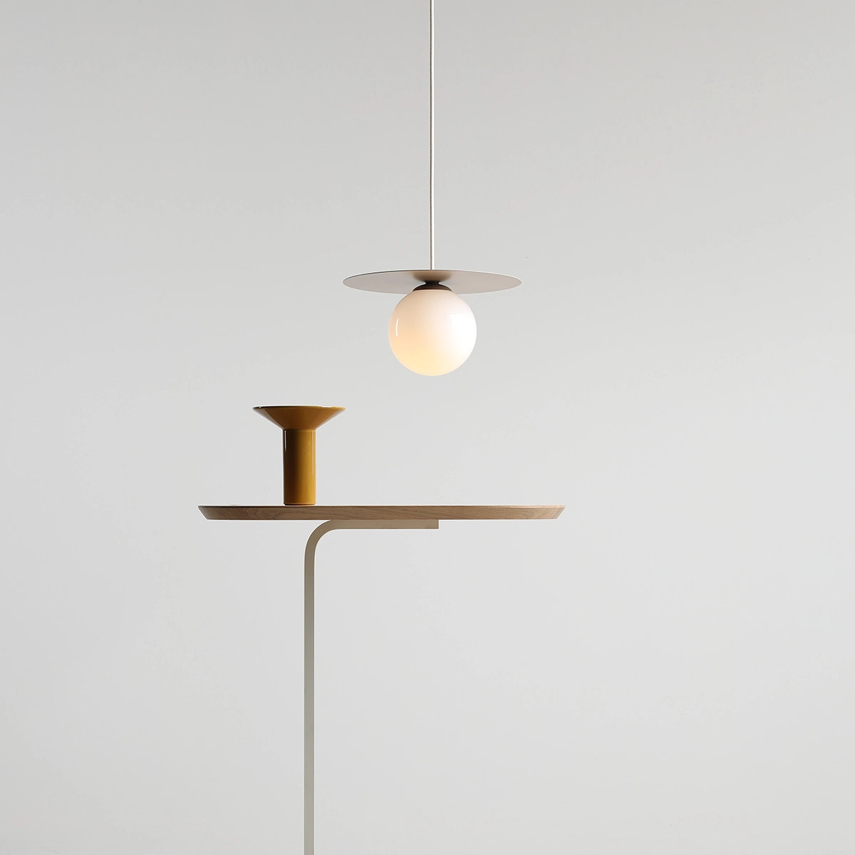 Lampa wisząca Loop Beige S z kloszem w kształcie kuli, zawieszona na cienkim, białym przewodzie. U podstawy znajduje się stolik z drewnianym blatem i żółtym, ceramicznym elementem. Tło jasne, minimalistyczne.
