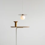 Lampa wisząca Loop Beige S z kloszem w kształcie kuli, zawieszona na cienkim, białym przewodzie. U podstawy znajduje się stolik z drewnianym blatem i żółtym, ceramicznym elementem. Tło jasne, minimalistyczne.