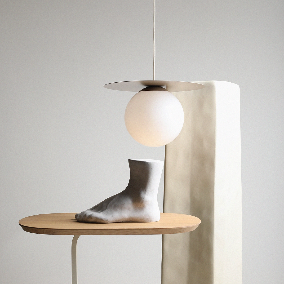 Lampa wisząca Loop Beige M z jednym źródłem światła E27, z kulistym, matowym kloszem i metalowym, okrągłym daszkiem, zawieszona nad rzeźbą stopy na drewnianym stole. Tło w jasnych odcieniach, minimalistyczna aranżacja.