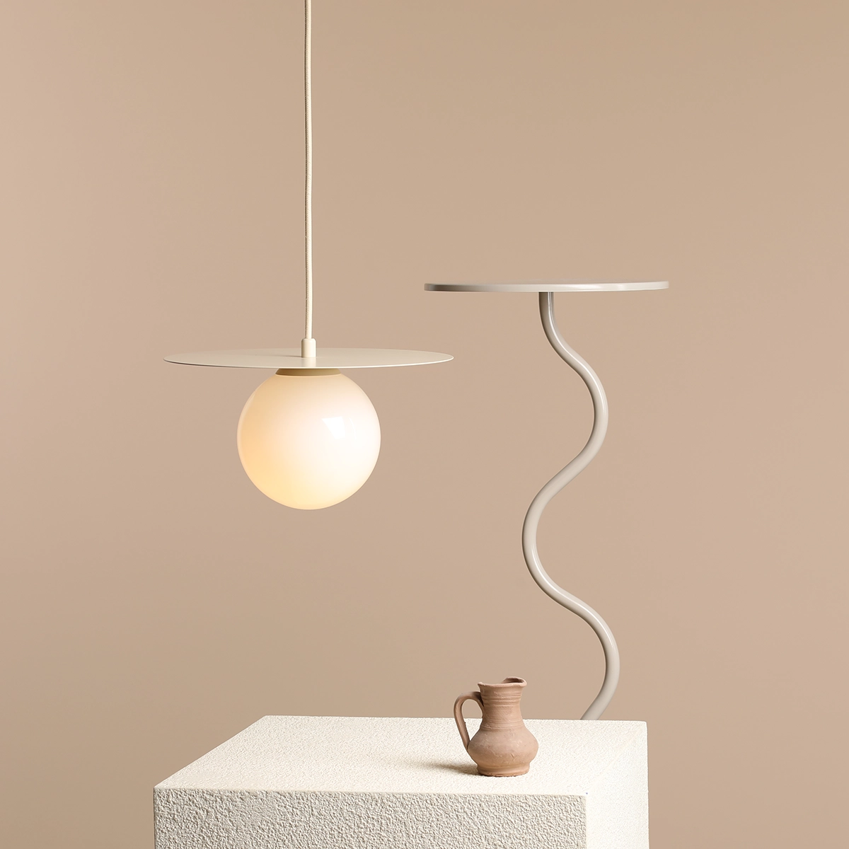 Lampa wisząca Loop Cream S z kloszem w kształcie kuli, w kolorze mlecznym, zawieszona na białym przewodzie. Okrągła, płaska podsufitka w jasnym kolorze. Obok znajduje się stół z falistym nogą oraz gliniana dzbanek na jasnym tle.