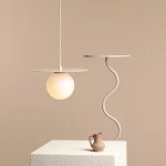 Lampa wisząca Loop Cream S z kloszem w kształcie kuli, w kolorze mlecznym, zawieszona na białym przewodzie. Okrągła, płaska podsufitka w jasnym kolorze. Obok znajduje się stół z falistym nogą oraz gliniana dzbanek na jasnym tle.