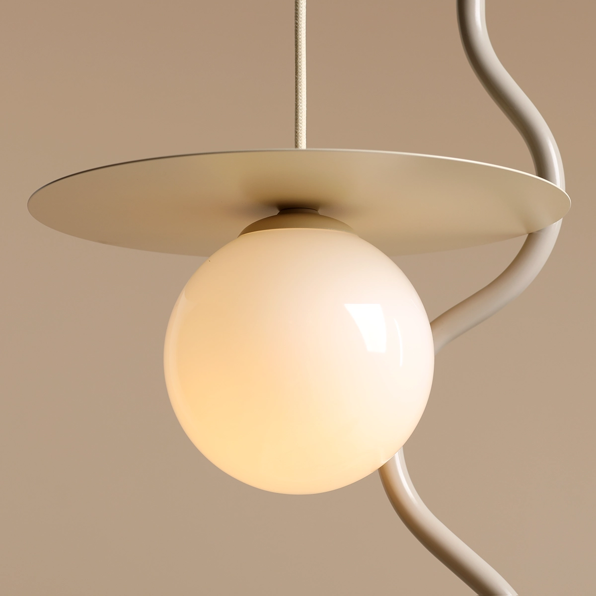 Lampa wisząca Loop Cream S z pojedynczym źródłem światła E14. Okrągła, matowa kula w kremowym kolorze zawieszona na prostokątnej, metalowej podstawie w jasnym odcieniu. Widoczne zakrzywione elementy mocujące. Tło w neutralnym kolorze.