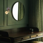 Kinkiet Paloma 1xE14 w kolorze mosiądzu z dymną kloszem, zamontowany na zielonej ścianie obok okrągłego lustra. W tle widoczne zasłony w odcieniu zieleń oraz drewniane biurko z filiżanką.