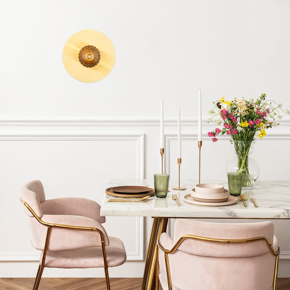 Mosiężny kinkiet Paloma z dymnym kloszem, umieszczony na ścianie, w otoczeniu eleganckiego stołu z różowymi krzesłami, świecami i wazonem z kwiatami. Kinkiet ma okrągły kształt z dekoracyjnym detalem w centrum.