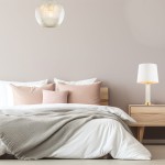 Lampa wisząca Paloma D25 w kolorze mosiądzu i szarości, o okrągłym kształcie z żebrowanym kloszem, zawieszona nad łóżkiem z poduszkami w odcieniach różu i bielą narzuty. W tle drewniana szafka nocna z lampką.