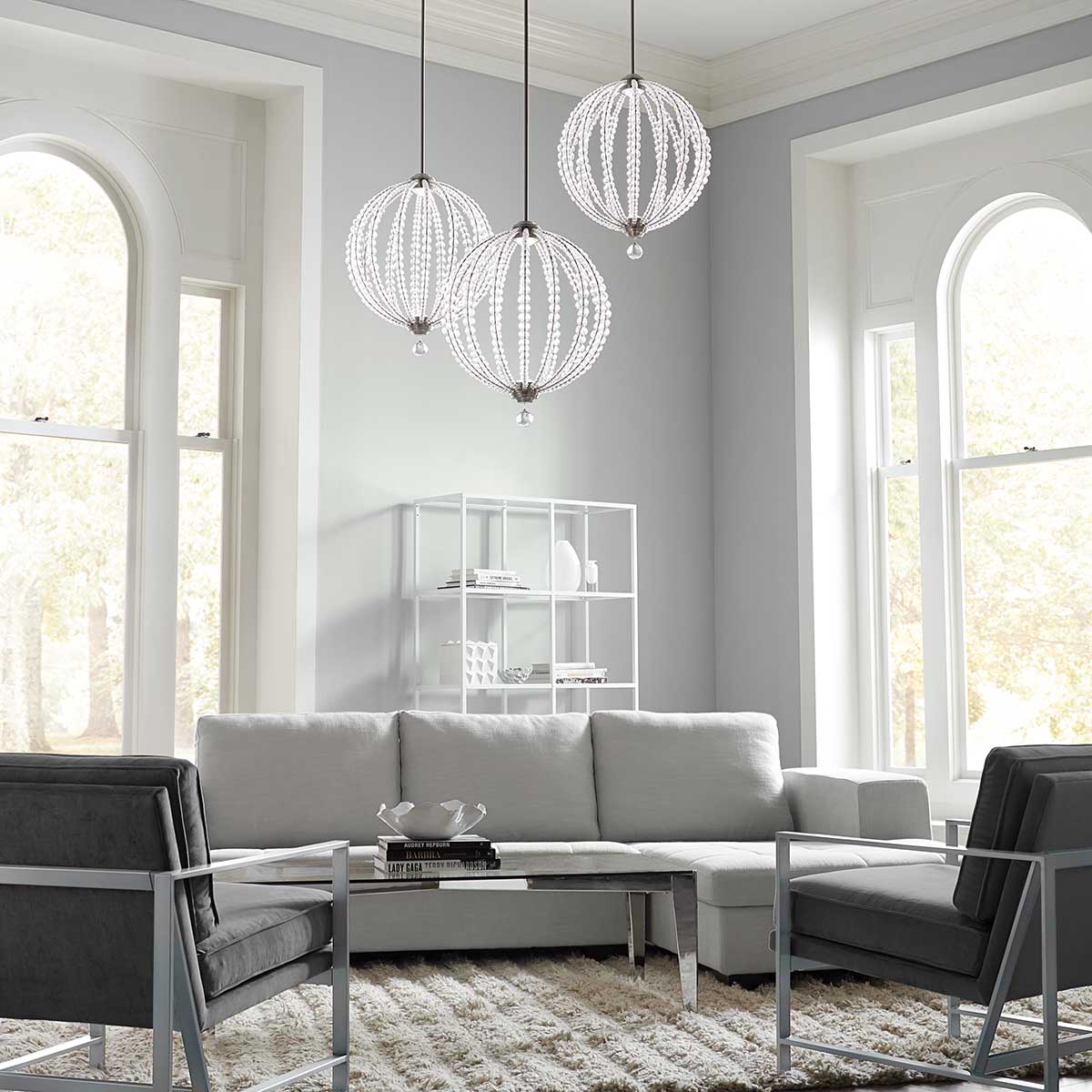 Lampa wisząca Oberlin L z niklu satynowego, z dwiema kulistymi oprawami LED, zawieszona w nowoczesnym wnętrzu. W tle widoczny szary narożnik, stolik oraz regał z białymi akcesoriami, na jasnej ścianie i w dużych oknach.