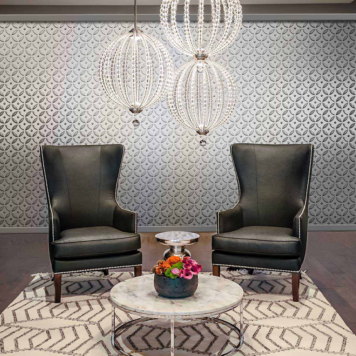 Lampa wisząca Oberlin M z dwoma kloszami LED o mocy 8W, wykonana z niklu satynowego, zawieszona nad stolikiem kawowym w eleganckim wnętrzu z czarnymi fotelami i dekoracyjnym dywanem. Tło w jasnych odcieniach z geometrycznym wzorem.
