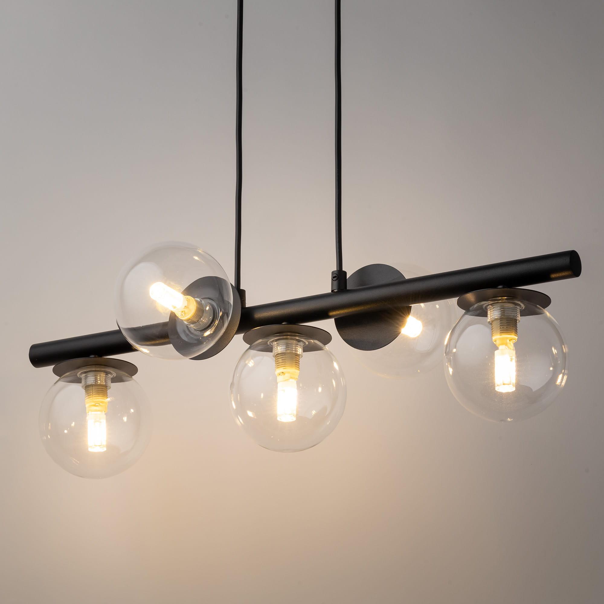 Lampa wisząca Avola z pięcioma źródłami światła G9. Czarna metalowa konstrukcja z transparentnymi szklanymi kloszami o okrągłym kształcie, zawieszona na dwóch przewodach. Stylowe połączenie nowoczesności z elegancją.