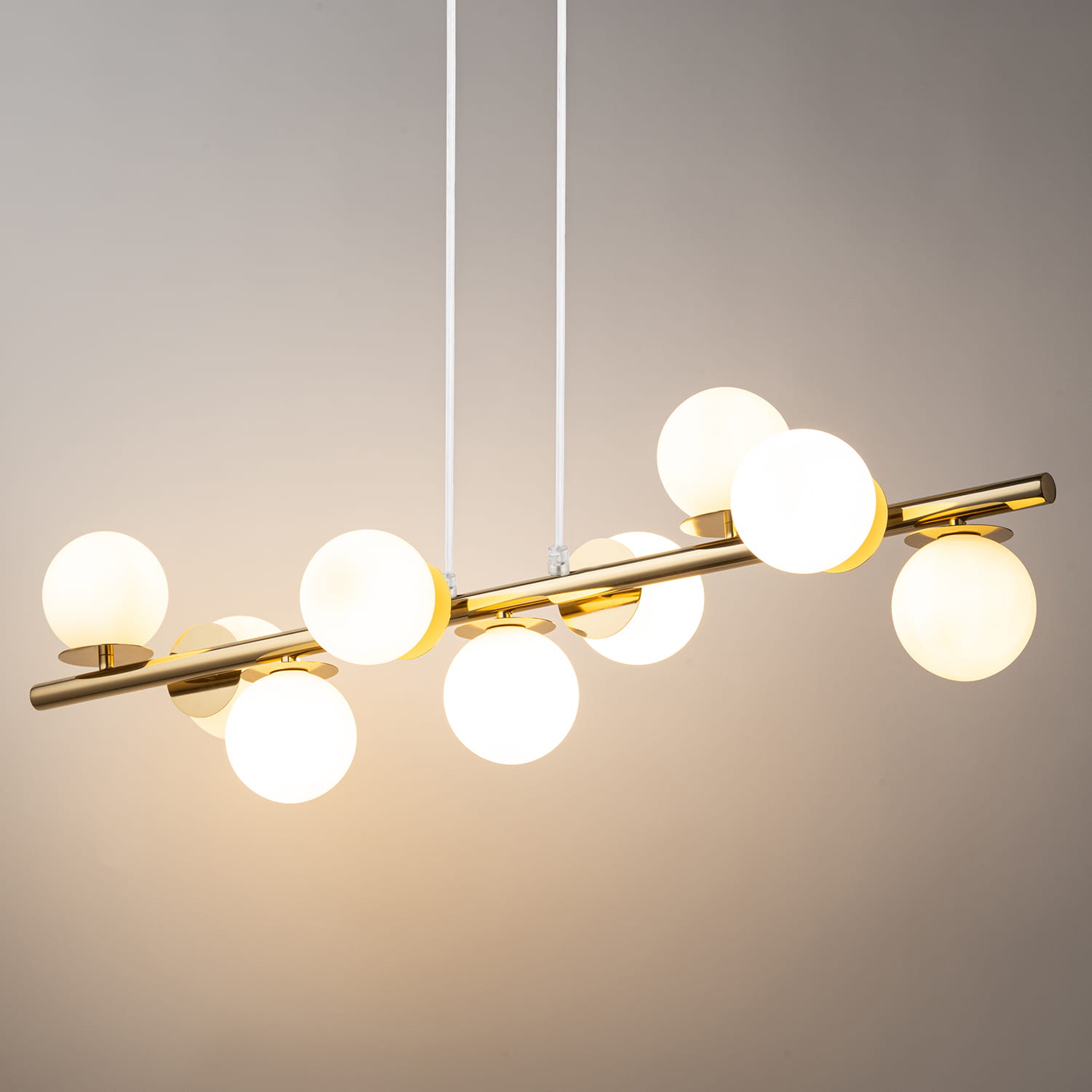 Lampa wisząca Avola z dziewięcioma kulistymi kloszami G9 w odcieniu białym, zawieszona na złotym metalowym stelażu. Klosze są równomiernie rozmieszczone, tworząc nowoczesny design. Dwa białe kable podtrzymują lampę. Tło w neutralnym kolorze.