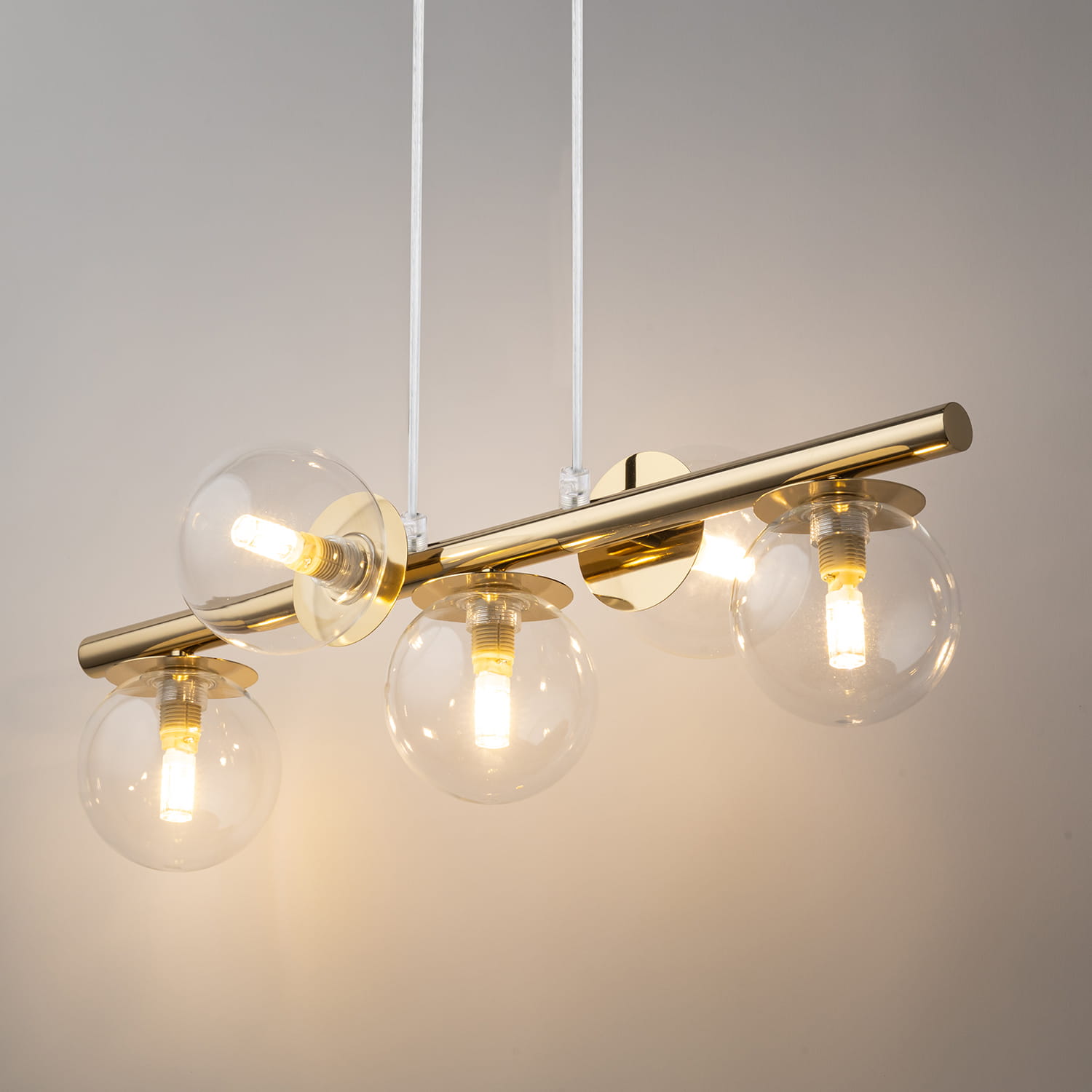 Lampa wisząca Avola z pięcioma kloszami G9 w złotym wykończeniu, z przezroczystymi, okrągłymi szkłami. Zawieszona na białych przewodach, umocowana na metalowej listwie. Klosze z widocznymi żarówkami emitującymi ciepłe światło.