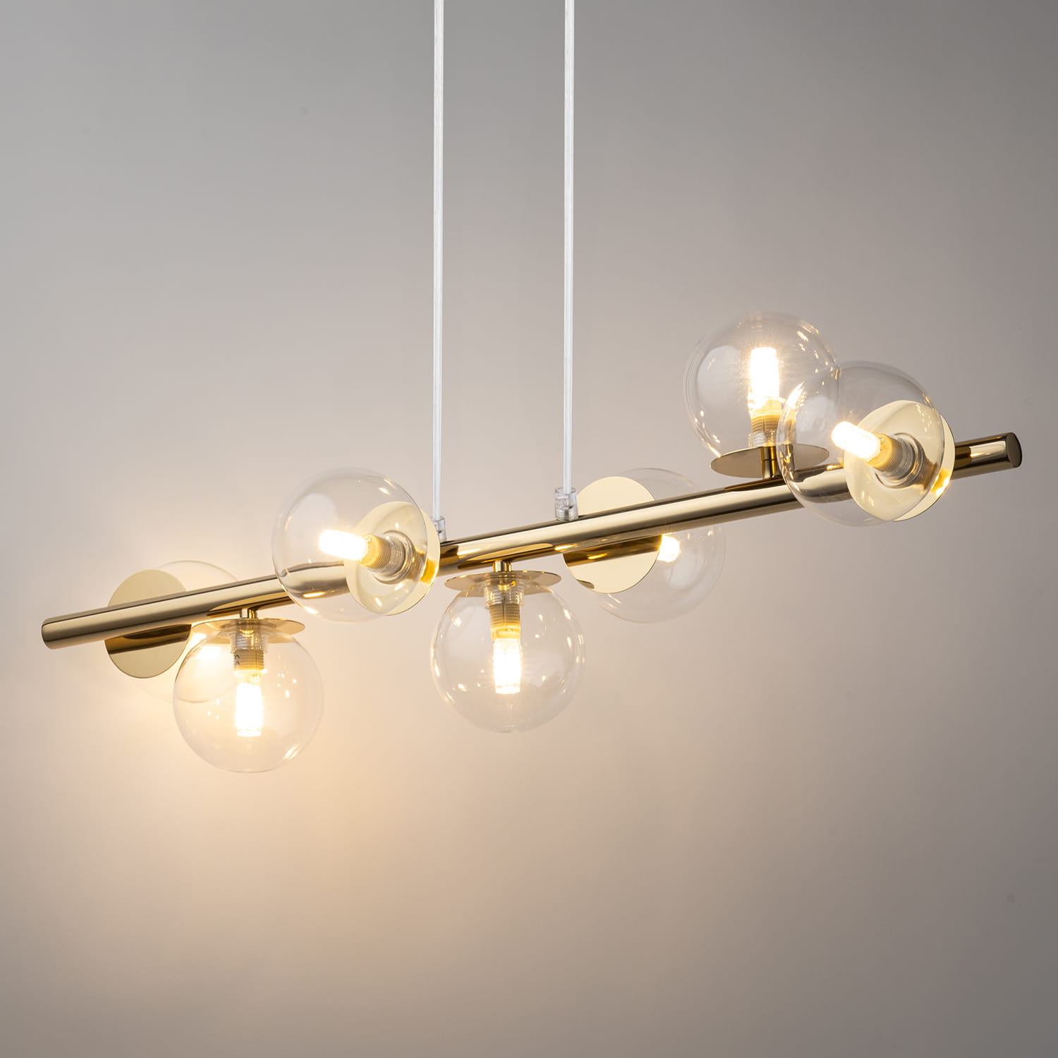 Lampa wisząca Avola z siedmioma źródłami światła G9, złożona z metalowego, złotego stelaża i przezroczystych, kulistych kloszy. Produkt zawieszony na dwóch białych przewodach, z lekkim podświetleniem w tle.