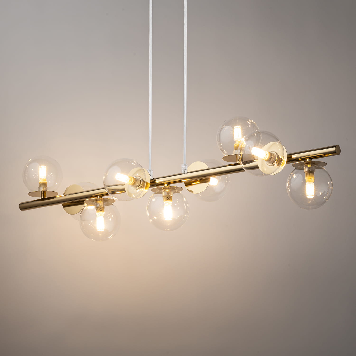 Lampa wisząca Avola w kolorze złotym z przezroczystymi kloszami, zawieszona na białych przewodach. Składa się z dziewięciu żarówek G9 umieszczonych na poziomej metalowej rurze, dając nowoczesny efekt świetlny.