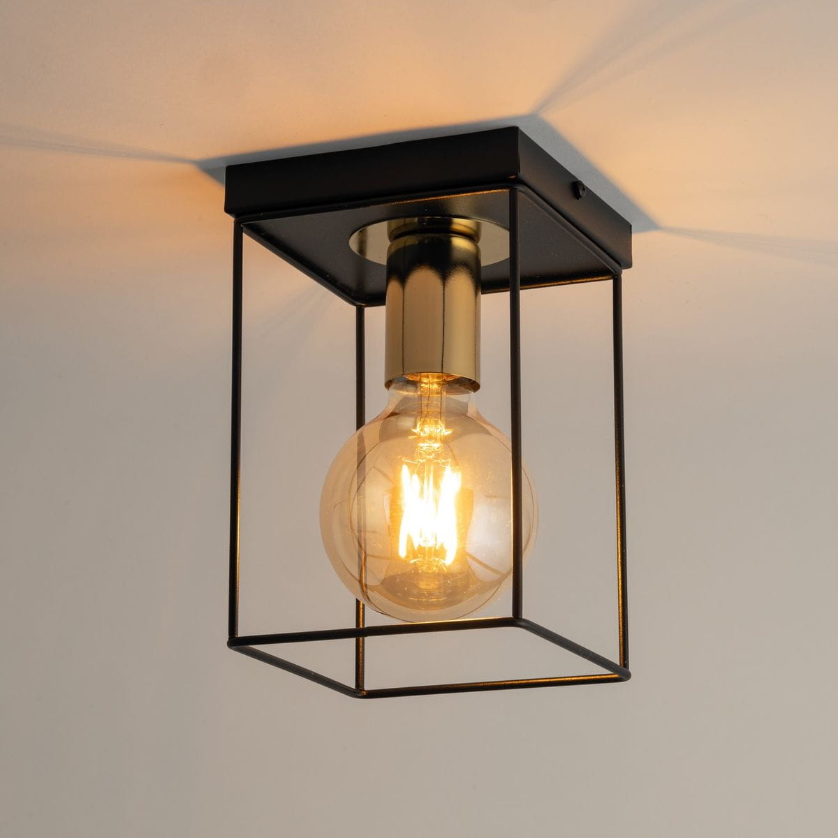 Czarna lampa sufitowa Vox z metalową klatką i złotym detalem, z lampą E27. Widoczny szklany klosz z żarówką w środku, zamontowana na suficie, z delikatnym oświetleniem w tle.