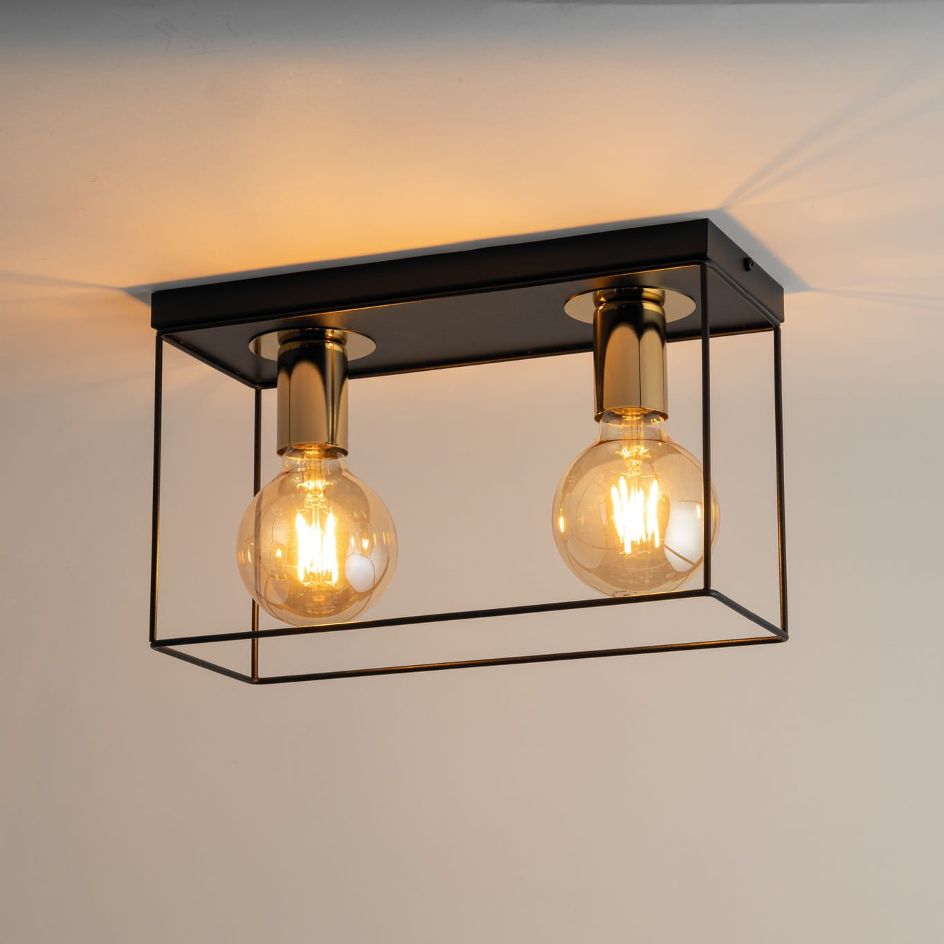 Czarna lampa sufitowa Vox z podwójnym źródłem światła E27, z metalową konstrukcją w złotym wykończeniu. Ujęcie z dołu pokazuje szklane żarówki, w prostokątnej ramie, zamontowana na białym suficie.