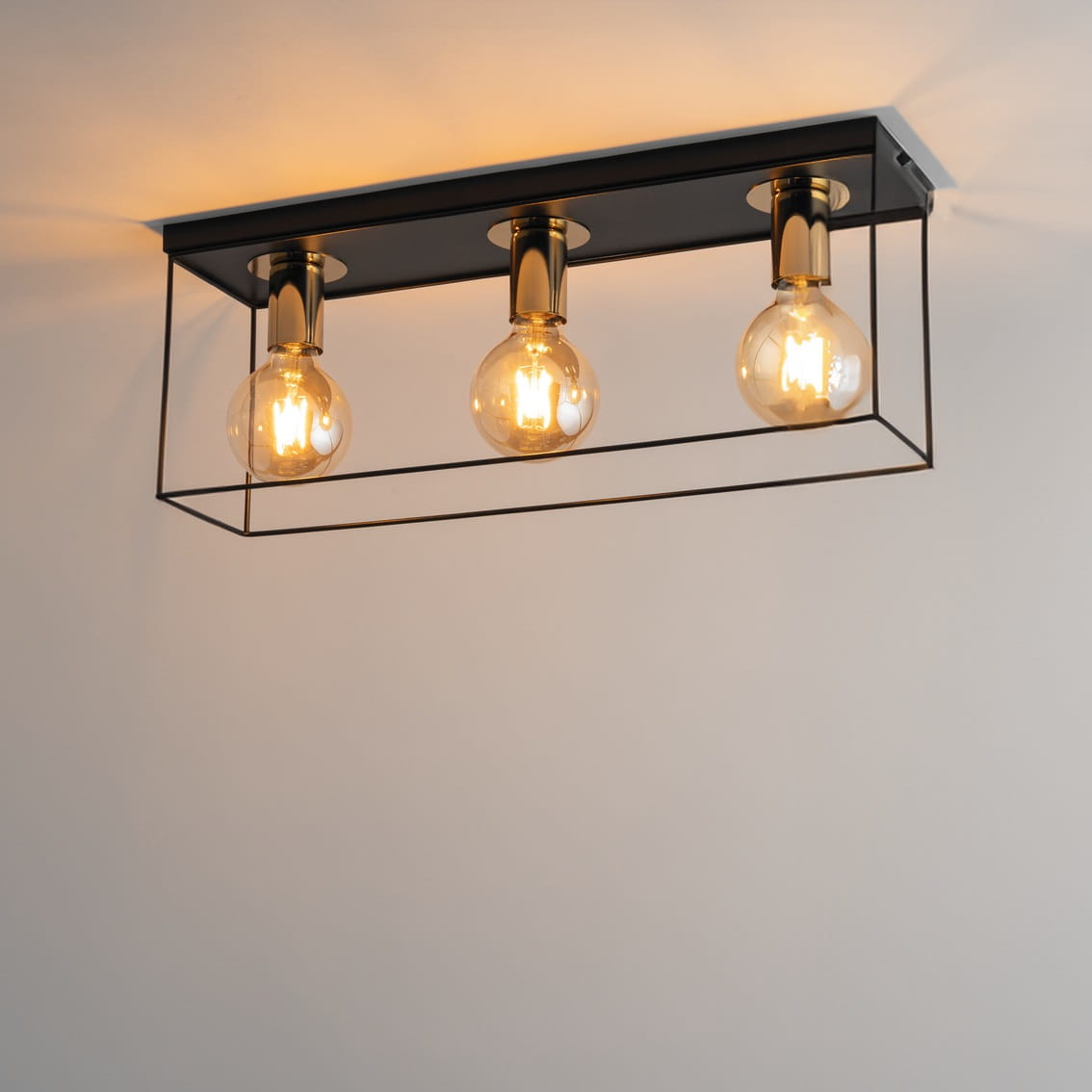 Lampa sufitowa Vox z trzema oprawami E27, w czarnej metalowej ramie z dekoracyjnymi złotymi wykończeniami. Ujęcie z góry, ukazujące przezroczyste żarówki, zawieszona na ścianie. Prosta, nowoczesna konstrukcja.