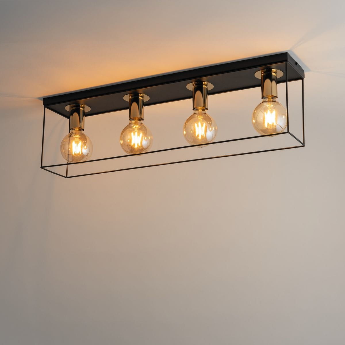 Czarna lampa sufitowa Vox z czterema oprawami E27, umieszczona w prostokątnej, metalowej ramie. Szklane żarówki w złotych mocowaniach emitują ciepłe światło, tworząc nowoczesny akcent w aranżacji wnętrza. Widok z boku, zamontowana na suficie.