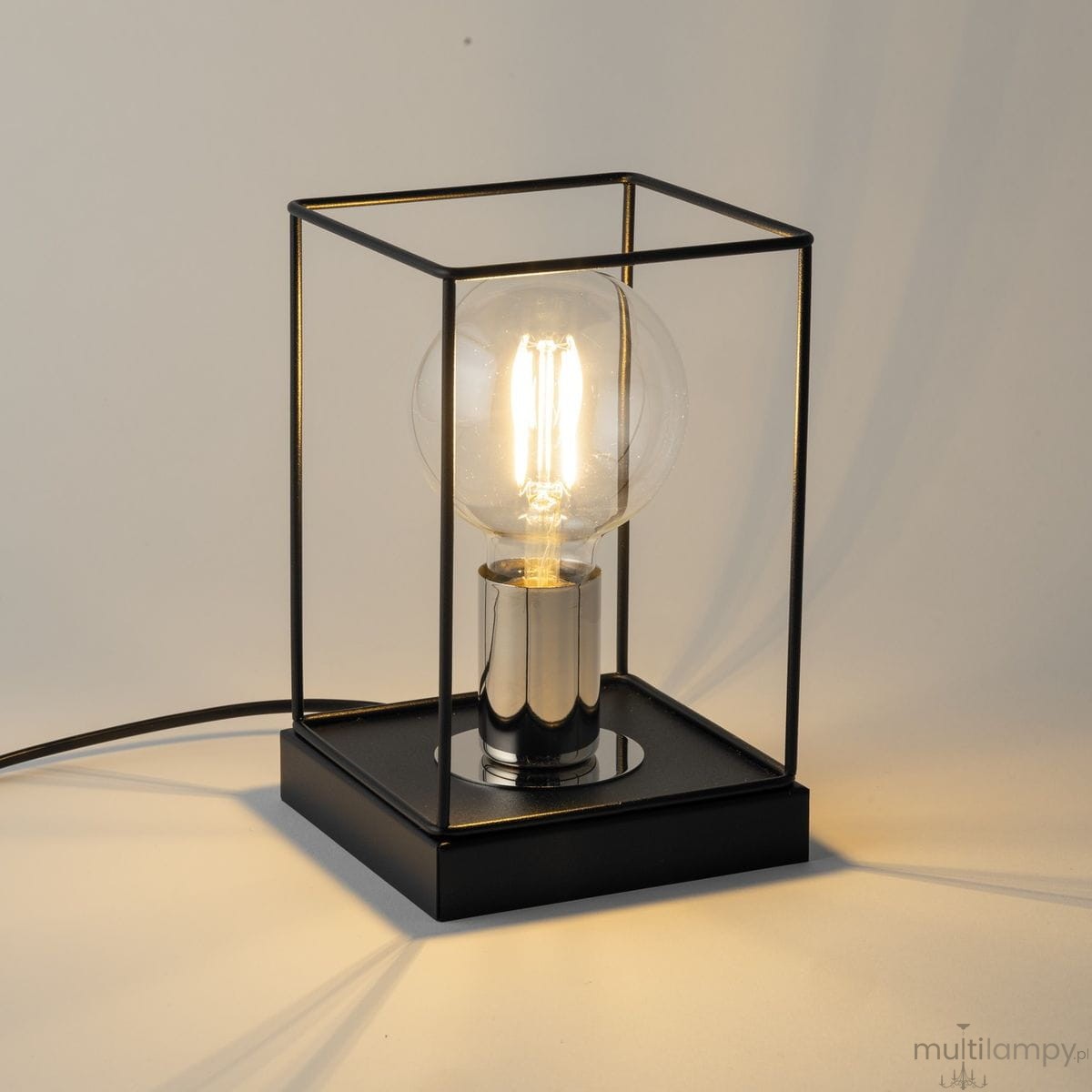 Lampka stołowa Vox 1xE27 w kolorze czarnym i srebrnym, z przezroczystą żarówką w metalowej oprawie. Produkt ma prostokątną podstawę, lekko otwartą konstrukcję oraz widoczny kabel. Ujęcie z boku, ukazujące detale i efekty świetlne.