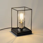 Lampka stołowa Vox 1xE27 w kolorze czarnym i srebrnym, z przezroczystą żarówką w metalowej oprawie. Produkt ma prostokątną podstawę, lekko otwartą konstrukcję oraz widoczny kabel. Ujęcie z boku, ukazujące detale i efekty świetlne.