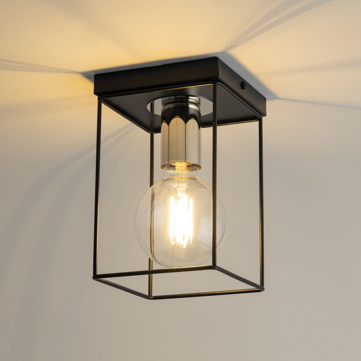 Lampa sufitowa Vox w kolorze czarnym i srebrnym, z metalową ramą w kształcie sześcianu. Widoczna żarówka E27 o okrągłym kształcie, emitująca ciepłe światło. Ujęcie z dołu, pokazujące detale konstrukcji i cień rzucany na sufit.