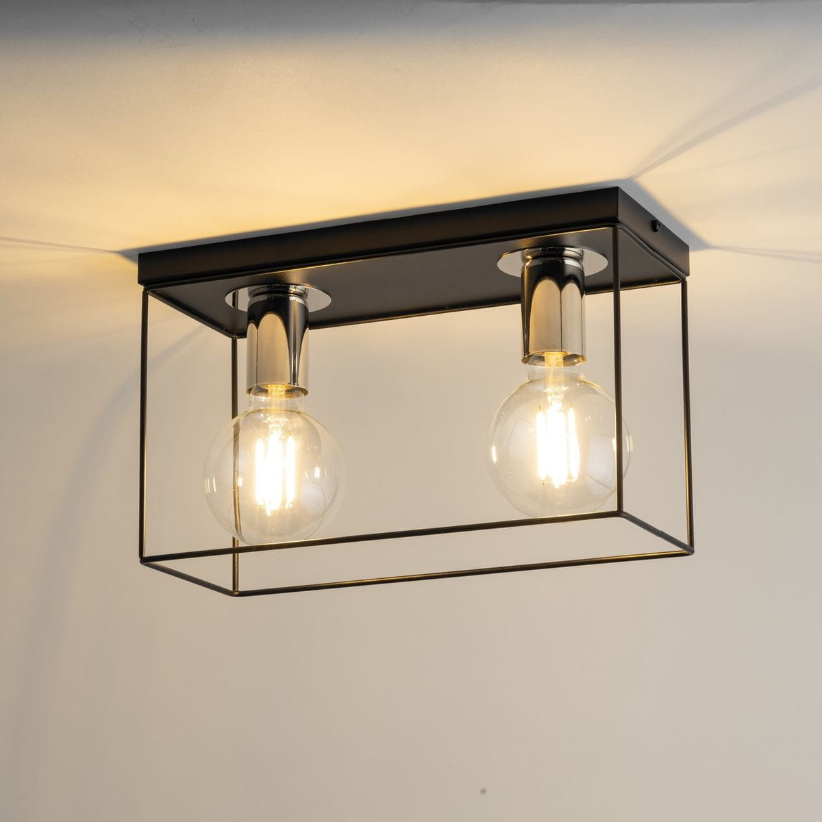 Lampa sufitowa Vox w kolorze czarnym i srebrnym z dwoma gniazdami E27. Posiada prostokątną, metalową konstrukcję z przezroczystymi żarówkami, zamontowana na suficie. Widoczny elegancki design z minimalistycznym wyglądem.