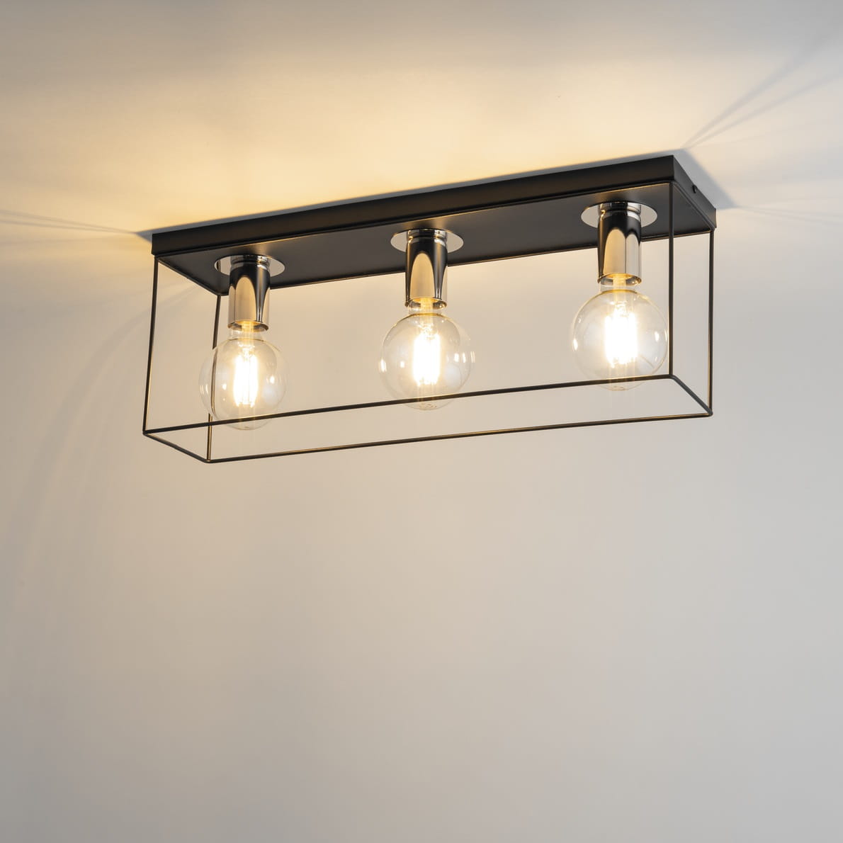 Lampa sufitowa Vox w kolorze czarnym i srebrnym z trzema oprawami na żarówki E27. Elementy metalowe w formie prostokątnej ramki, w której umieszczone są przezroczyste żarówki. Ujęcie z góry, ukazujące montaż na suficie.