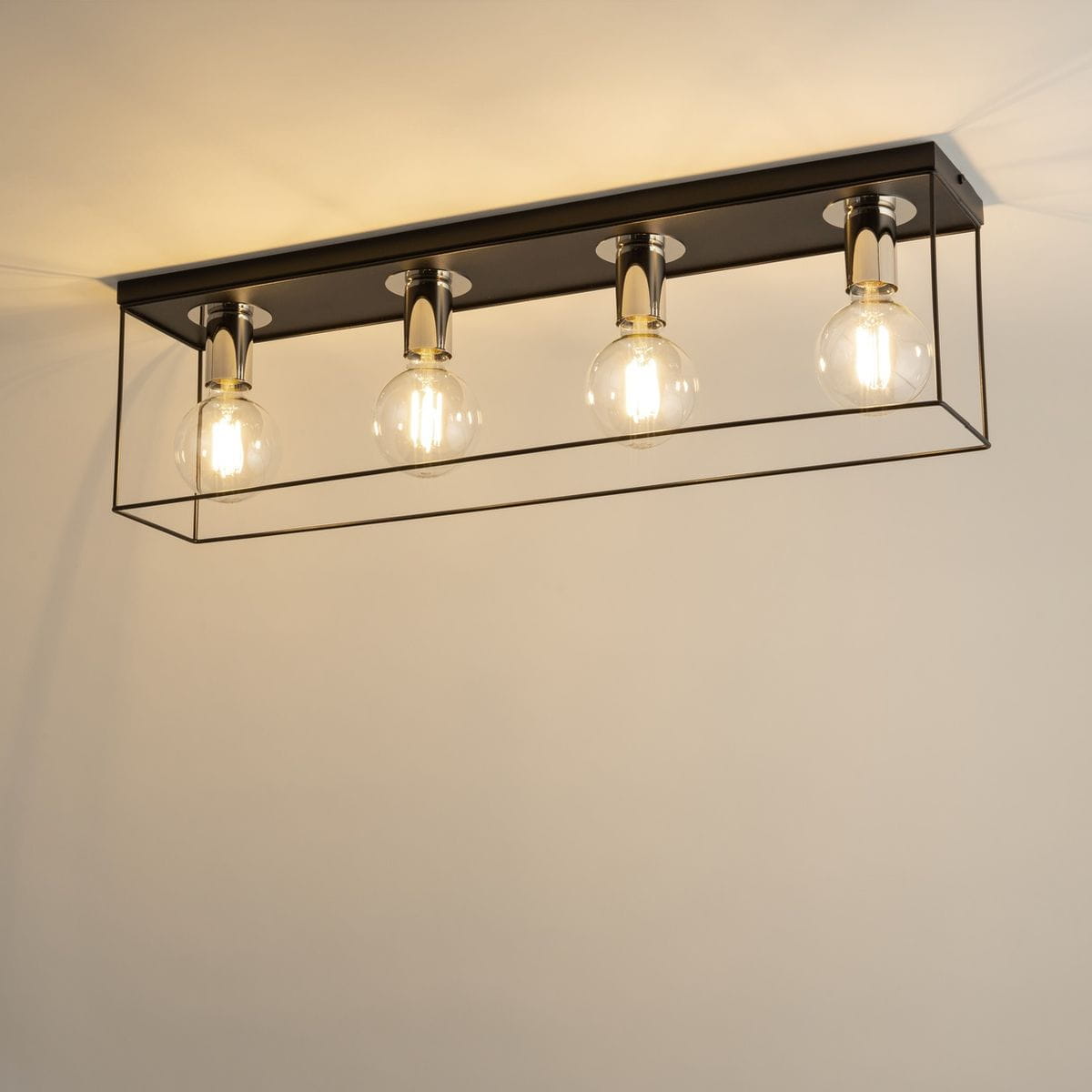Czarna lampa sufitowa Vox z czterema oprawami E27, z metalową ramą w kolorze srebrnym. Ujęcie z góry, z widocznymi przezroczystymi żarówkami. Lampa ma prostokątny kształt i minimalistyczny design, zawieszona na jasnym tle.