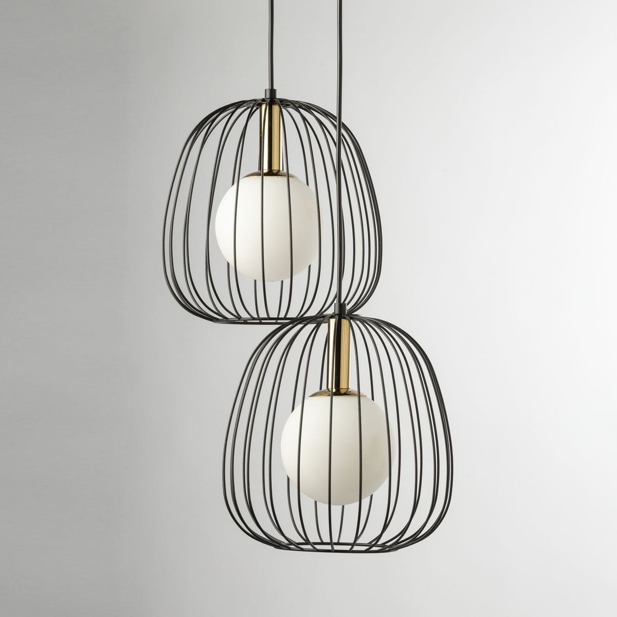 Lampa wisząca Bell z dwoma oprawami G9, w kolorze czarnym i złotym. Posiada metalową klatkę w kształcie owalnym, w której znajdują się białe, okrągłe klosze. Zawieszona na cienkich linkach, prezentuje nowoczesny design. Tło neutralne, jasne.