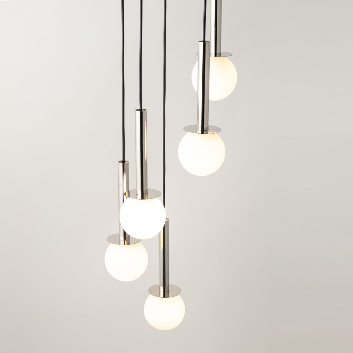 Lampa wisząca Daisy, pięć źródeł światła G9, srebrne metalowe elementy oraz białe kuliste klosze. Produkt zawieszony na czarnych przewodach, w górnej części widoczna metalowa podstawa, kształt cylindryczny. Tło jasne, jednolite.
