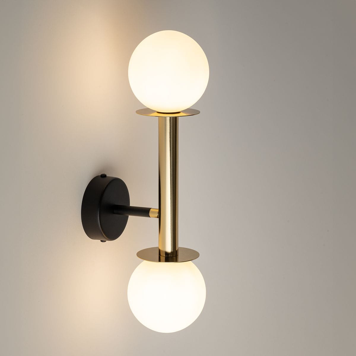 Kinkiet Daisy w kolorze złotym i białym z dwiema kulistymi lampami. Metalowa konstrukcja z gładkim wykończeniem, zamontowana na czarnej podstawie. Światło emitowane przez białe klosze tworzy subtelny efekt oświetlenia.