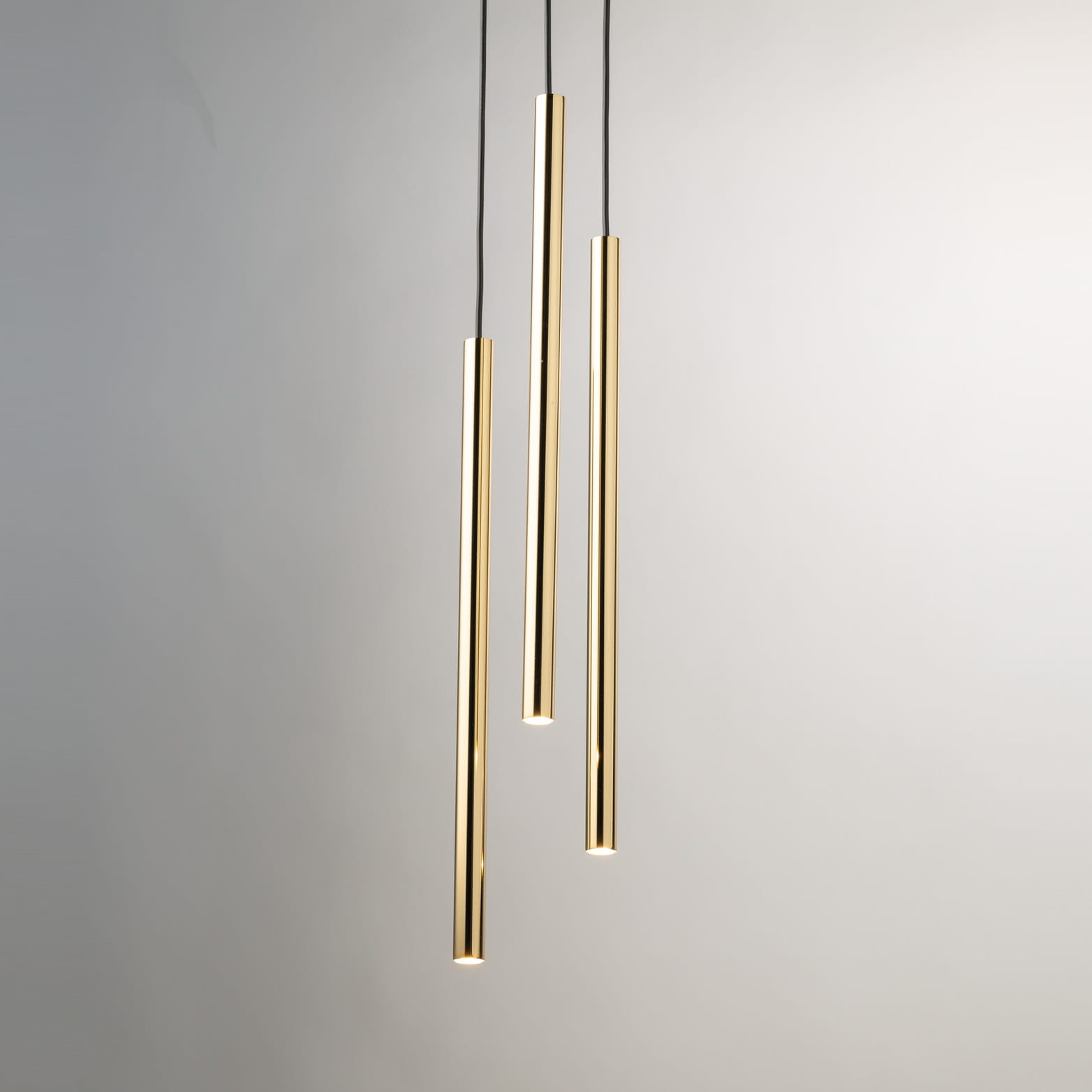 Lampa wisząca Sopel Lux w kolorze czarnym i złotym, składająca się z trzech cienkich, cylindrycznych elementów. Zawieszona na czarnych linkach, emitująca światło z dolnej części, na jasnym tle.