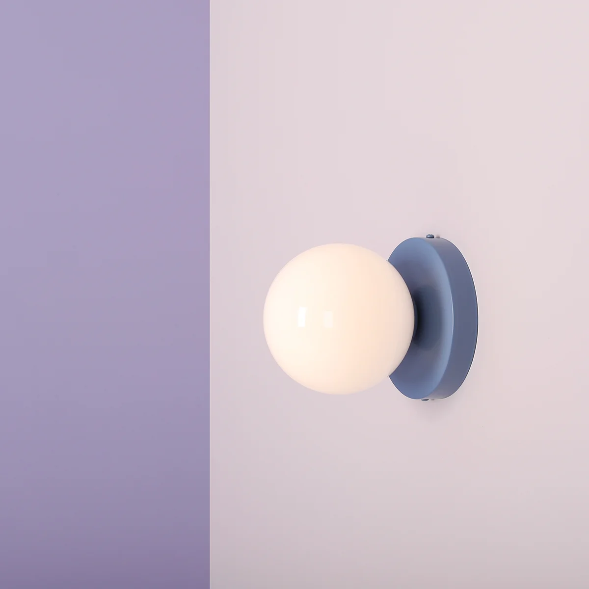 Kinkiet Ball II S w kolorze dusty blue z okrągłą, białą, matową kulą. Zamontowany na ścianie, tworzy minimalistyczny i nowoczesny akcent. Ściana w tle w jasnym odcieniu fioletu i różu.