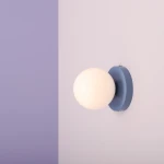 Kinkiet Ball II S w kolorze dusty blue z okrągłą, białą, matową kulą. Zamontowany na ścianie, tworzy minimalistyczny i nowoczesny akcent. Ściana w tle w jasnym odcieniu fioletu i różu.