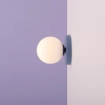 Kinkiet Ball II S w kolorze Dusty Blue, z okrągłą, białą szklaną kulą, zamontowany na ścianie. Ujęcie z boku, na tle w kolorze fioletowym. Metalowa oprawa w delikatnym niebieskim odcieniu.