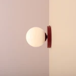 Kinkiet Ball II S w kolorze czerwonego wina z białą, okrągłą oprawą. Zamontowany na ścianie, w perspektywie bocznej, ukazuje gładką powierzchnię oraz minimalistyczny design. Idealny do nowoczesnych wnętrz.