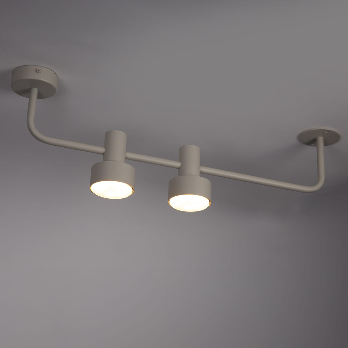 Lampa sufitowa Mona w kolorze szarym/beżowym, z dwiema oprawami na żarówki GX53. Ułożona poziomo, z widocznym metalowym ramieniem i okrągłymi kloszami, montowana do sufitu. Prosty design, odpowiednia do nowoczesnych wnętrz.