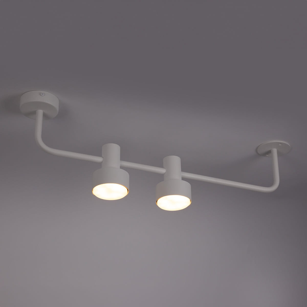 Lampa sufitowa Mona w białym kolorze z dwoma oprawami na żarówki GX53, zamontowana na suficie. Prosty, minimalistyczny design z metalowym stelażem. Kąt ujęcia z góry, ukazujący oba źródła światła.