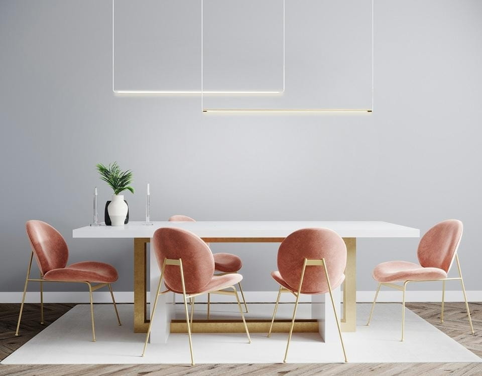 LED lampa wisząca Ombre Level 100 w kolorze złotym, z minimalistycznym, prostokątnym kształtem. Umieszczona nad białym stołem, w nowoczesnej aranżacji z różowymi krzesłami i rośliną w wazonie. Tło w stonowanym odcieniu szarości.