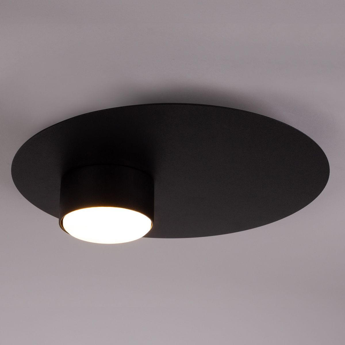 Czarna lampa sufitowa Star z jedną oprawą na żarówkę GX53. Okrągła, matowa podstawa z zamontowanym cylindrycznym kloszem, emitującym ciepłe światło. Ujęcie z góry, ukazujące minimalistyczny design.