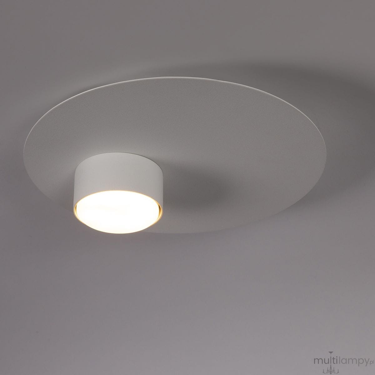 Biała lampa sufitowa Star z pojedynczym źródłem światła GX53. Okrągła, płaska osłona z matowego materiału z centralnym cylindrycznym elementem. Kąt ujęcia z góry, na jednolitym tle.