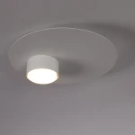 Biała lampa sufitowa Star z pojedynczym źródłem światła GX53. Okrągła, płaska osłona z matowego materiału z centralnym cylindrycznym elementem. Kąt ujęcia z góry, na jednolitym tle.