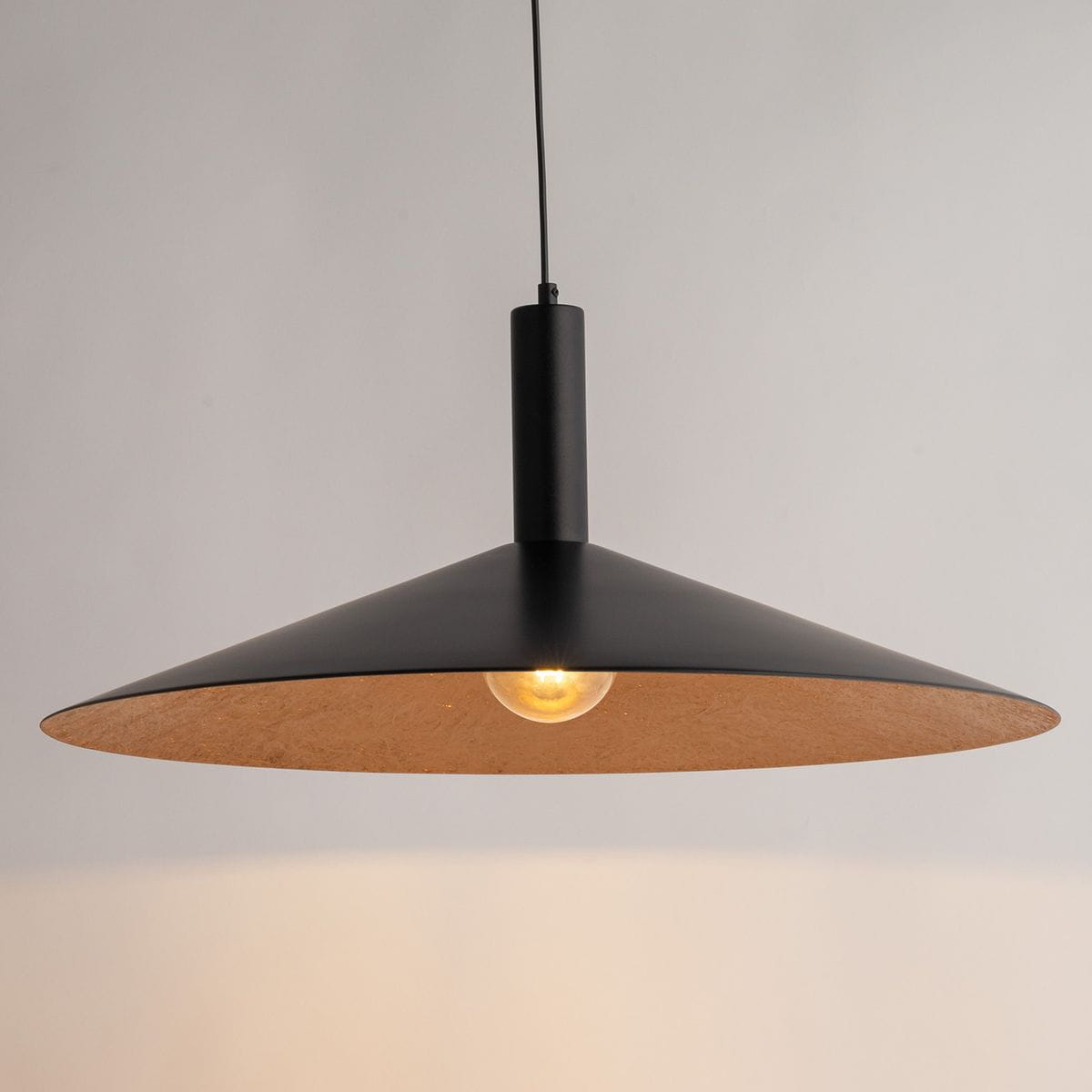 Czarna lampa wisząca Capri D70 z miedzianym wnętrzem, z pojedynczym źródłem światła E27. Elegancki, stożkowy kształt z matowego metalu, zawieszona na cienkim przewodzie w prostym otoczeniu.