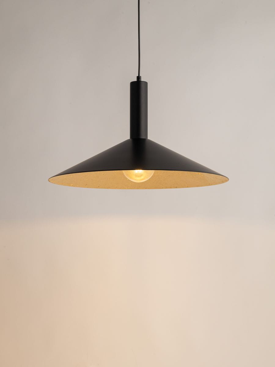 Lampa wisząca Capri D50 w kolorze czarnym z złotym wnętrzem, o stożkowym kształcie. Zawieszona na cienkim przewodzie, z oprawą na żarówkę E27. Widoczna żarówka emituje ciepłe światło, tło w jasnym odcieniu.