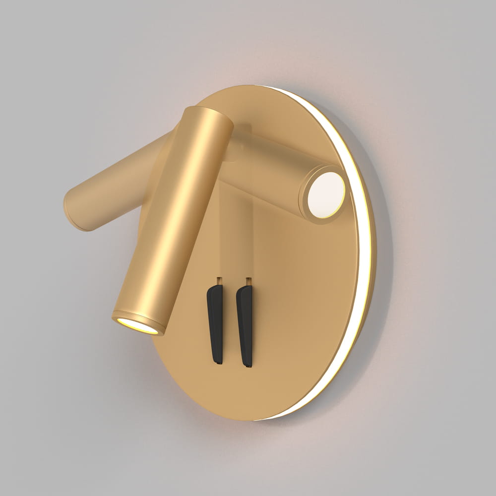 Kinkiet LED Ios 176, 2-punktowy, złoty, z okrągłą podstawą. Dwa ruchome ramiona z lampami w kształcie cylindrów, podświetlenie zewnętrzne. Elementy montażowe w czerni. Produkt zamontowany na gładkiej, jasnej ścianie.