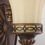 Kinkiet Drawing Room 1xE27 w postarzanym brązie. Okrągła podstawa z dekoracyjnym wzorem, z kloszem w kremowym kolorze, zdobionym metalową obręczą. Ujęcie z boku, eksponujące detale konstrukcji i fakturę materiałów.