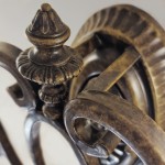 Kinkiet Drawing Room w postarzanym brązie, z detalami w formie dekoracyjnych, obłych kształtów. Produkt z dwiema oprawami na żarówki E27, widoczny z góry, z ozdobnym elementem na szczycie. Wykonany z metalu, nadaje się do wnętrz w stylu vintage.