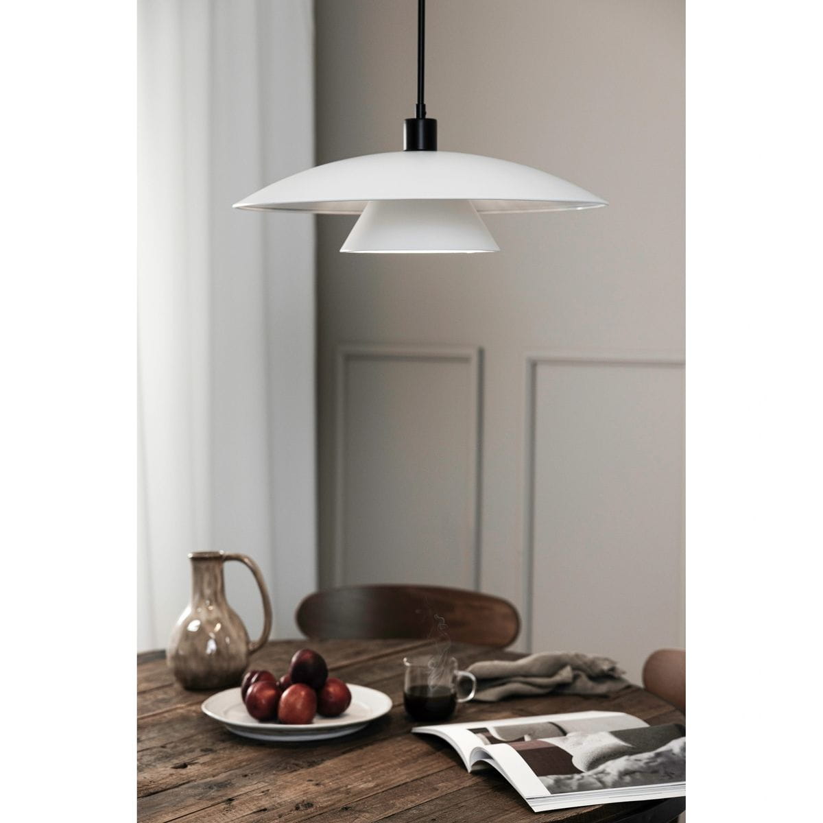 Lampa wisząca Millinge 1xE27 w kolorze czarnym i białym, z szerokim kloszem w kształcie stożka, zawieszona na długim przewodzie. W tle drewniany stół z filiżanką kawy, talerzem owoców i gazetą, w jasnym, minimalistycznym wnętrzu.